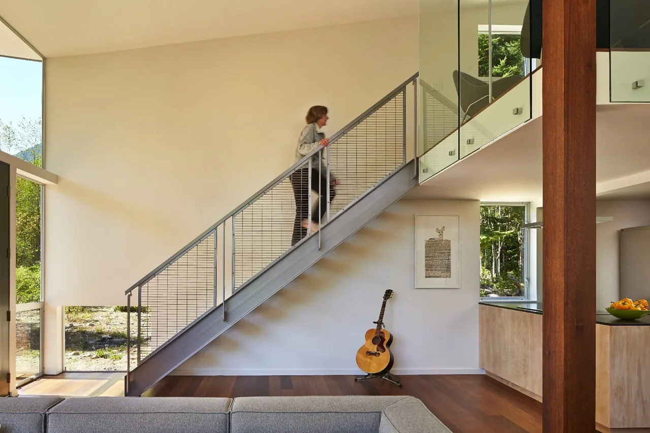 Modern living room with a metal railing staircase and large windows overlooking a forest setting.
