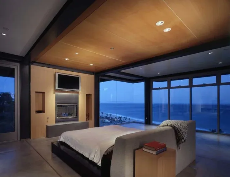 The Grenola Malibu residence bedroom features floor-to-ceiling glazing with ocean views, a fireplace, and a wooden ceiling.