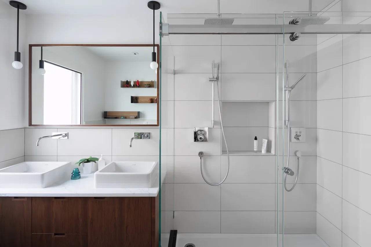 Bathroom featuring dual wall-mounted faucets, a large mirror, wooden vanity, and a glass shower enclosure with dual shower heads.