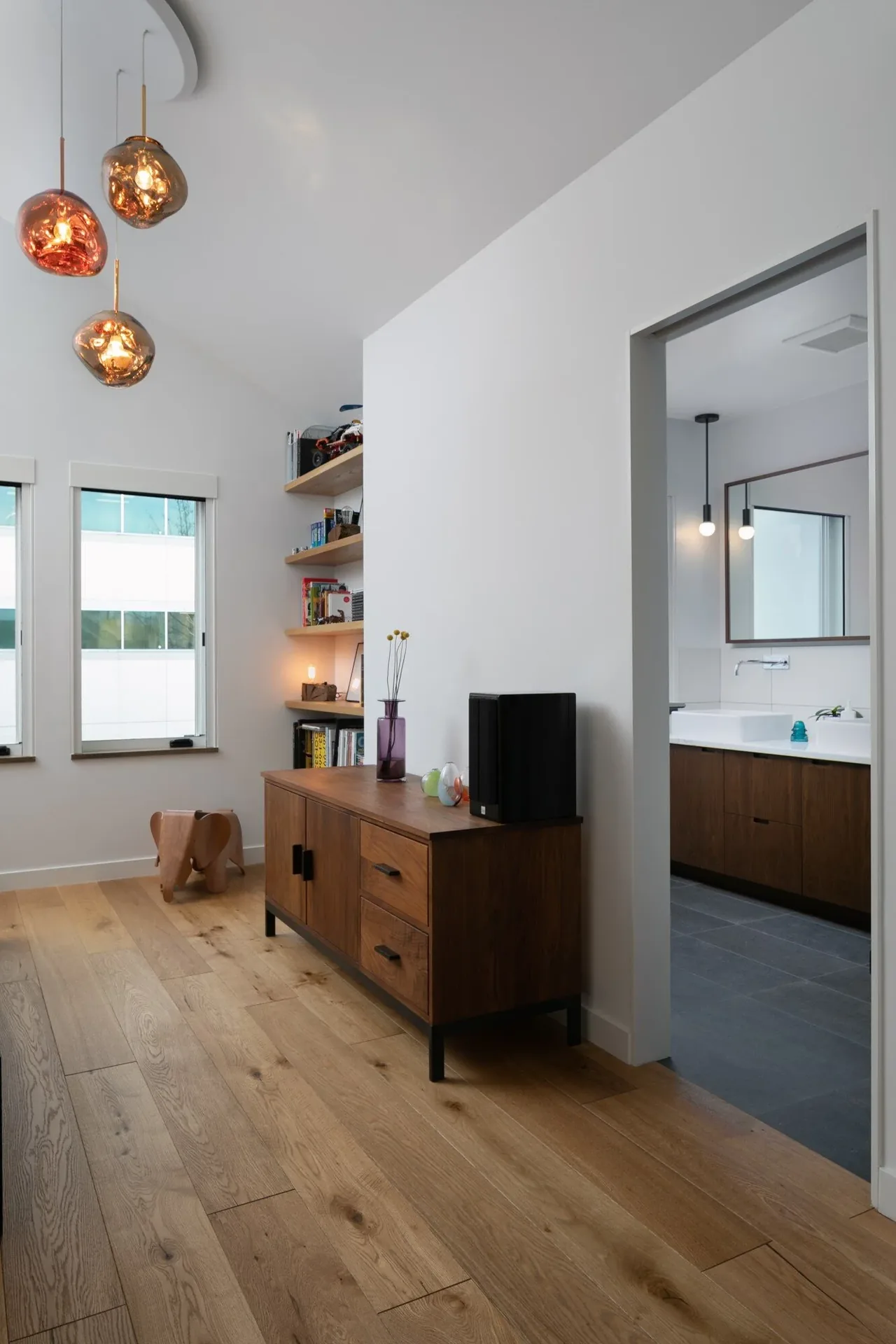 View of a hallway featuring wood flooring, a wooden sideboard, floating shelves, and an adjacent bathroom with pendant lights and a large mirror.