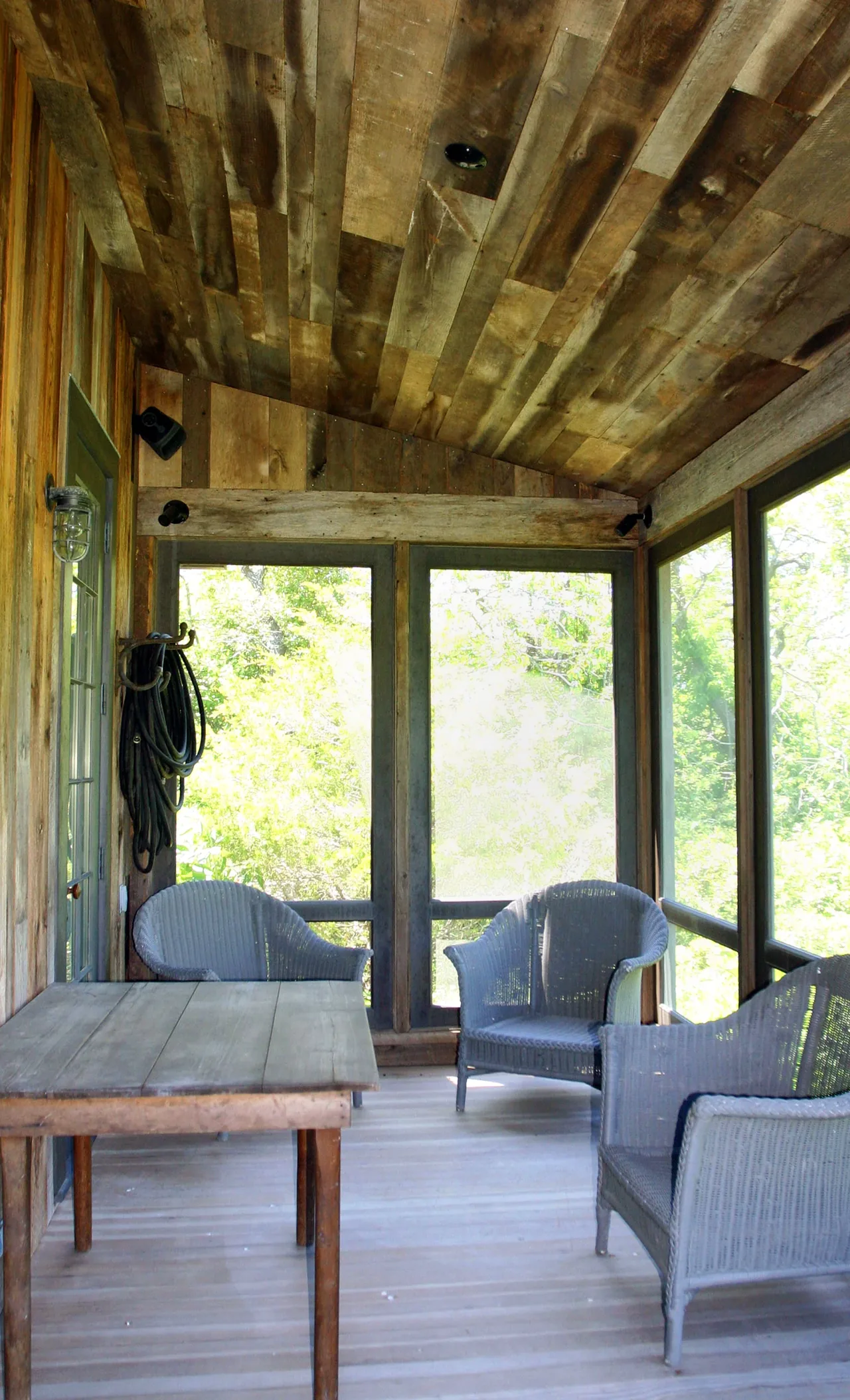 Rustic screened porch with wood plank ceiling and siding, furnished with a wooden table and wicker chairs.