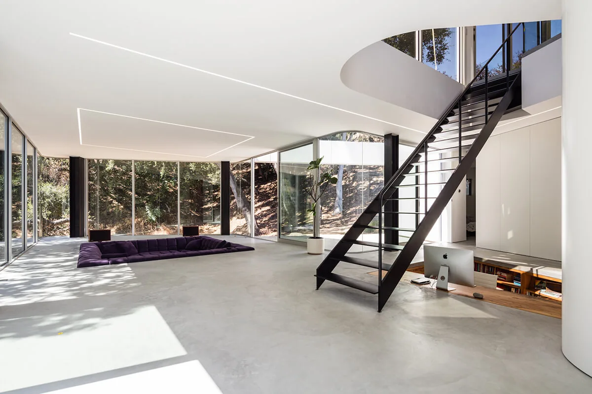 Living room with large glass walls looking out to woodland and a metal staircase leading to an upper floor.