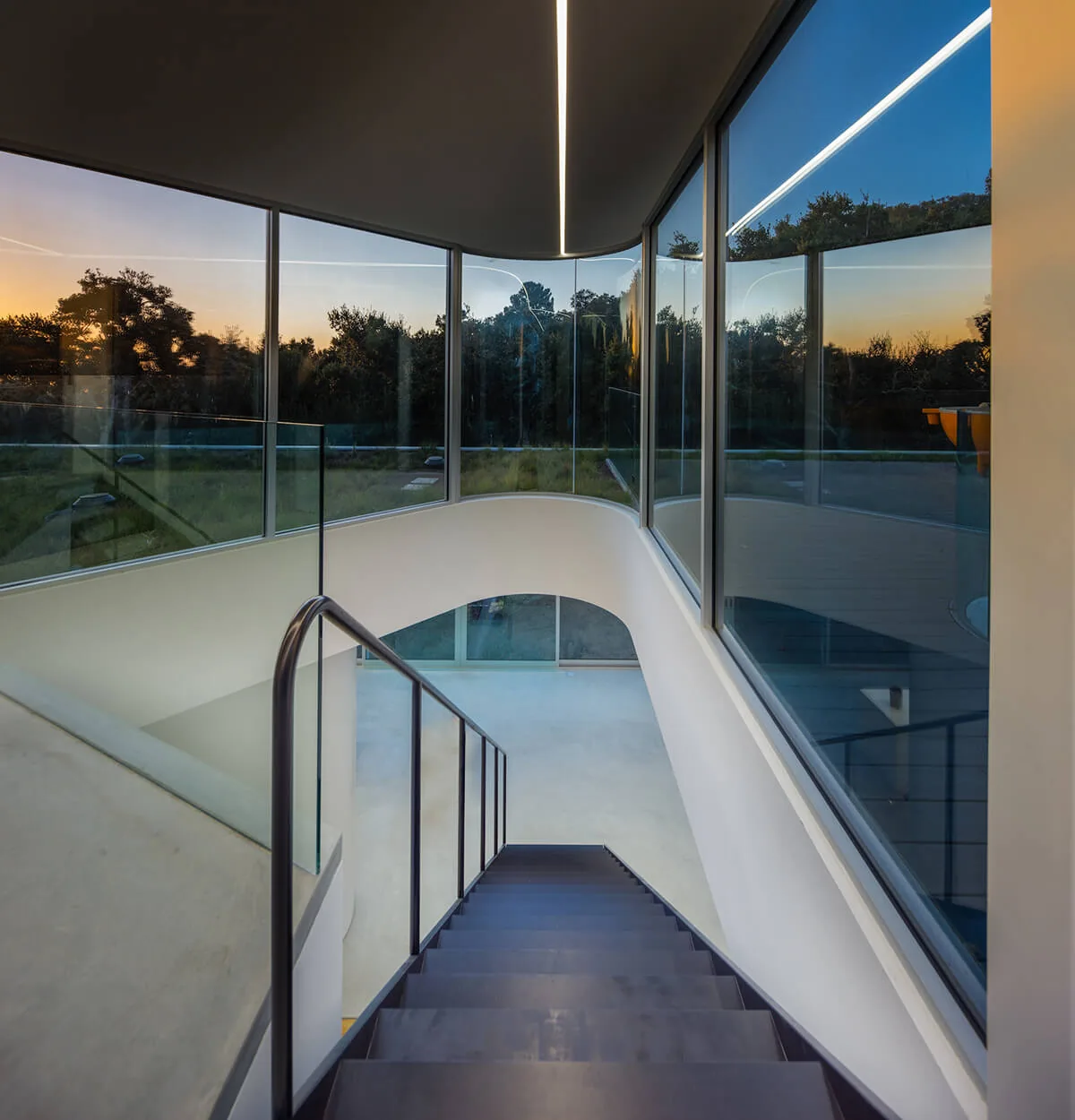 Curved staircase with glass railing and floor-to-ceiling windows overlooks wooded landscape during sunset.