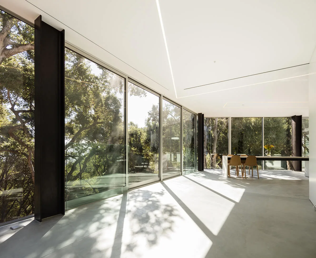 Open dining space with floor-to-ceiling glazing and black window frames overlooks a wooded landscape.