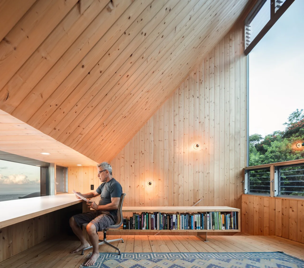 Wood-paneled study with sloped ceiling, built-in desk, floating bookshelf, and large windows showing outdoor trees and sky.