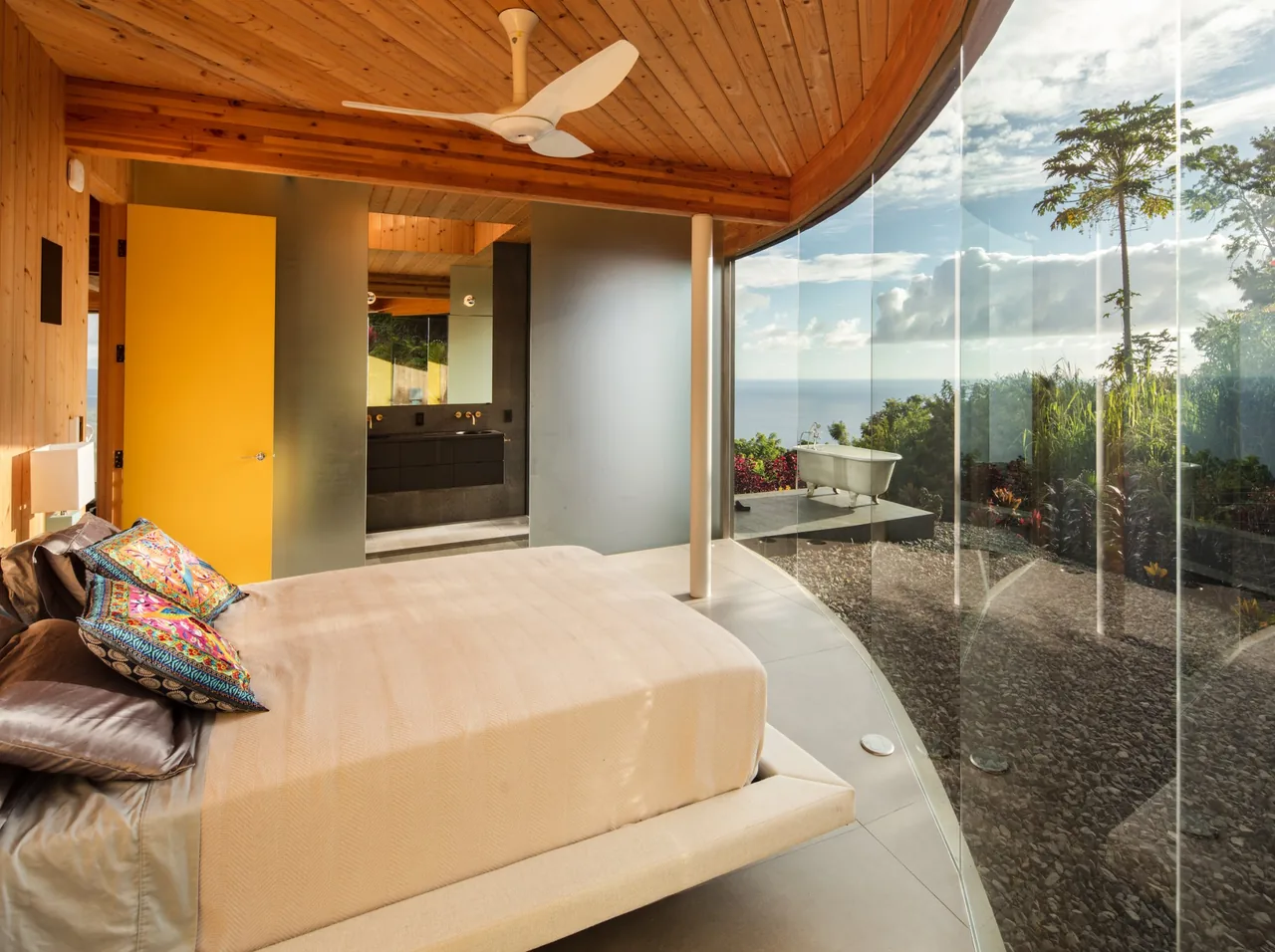 Bedroom in D Fin House features a curved glass wall overlooking outdoor landscaping and a freestanding tub on the patio.