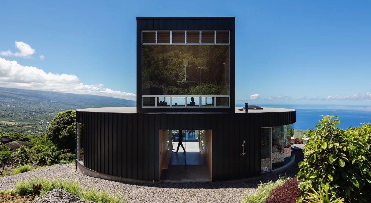 The D Fin House features a circular design with standing seam metal siding and a large square upper volume with expansive glazing.