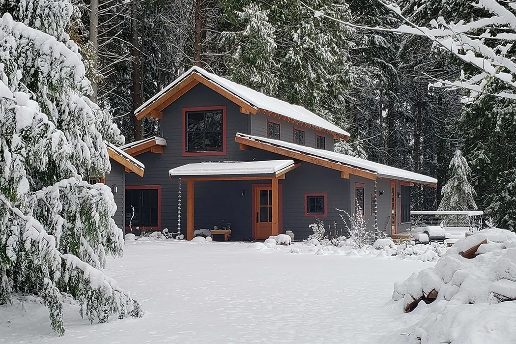 Snowy view of Bainbridge Island Farmhouse featuring dark siding, exposed wood beams, and a covered front porch.