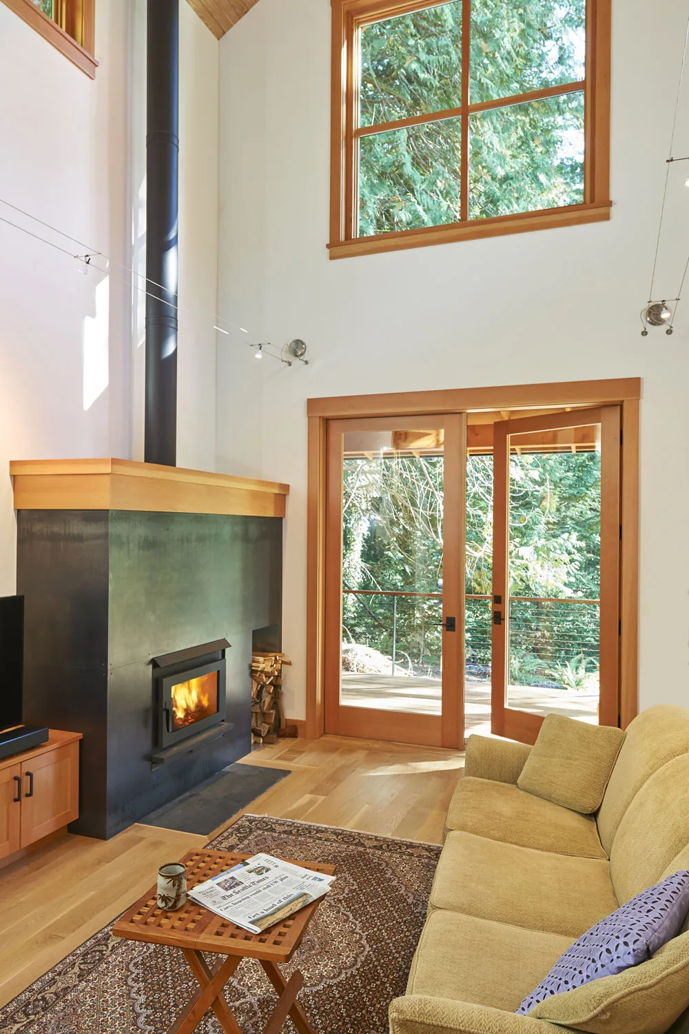 Living room of Bainbridge Island Farmhouse features a modern wood stove and large wooden framed doors and window with forest views.