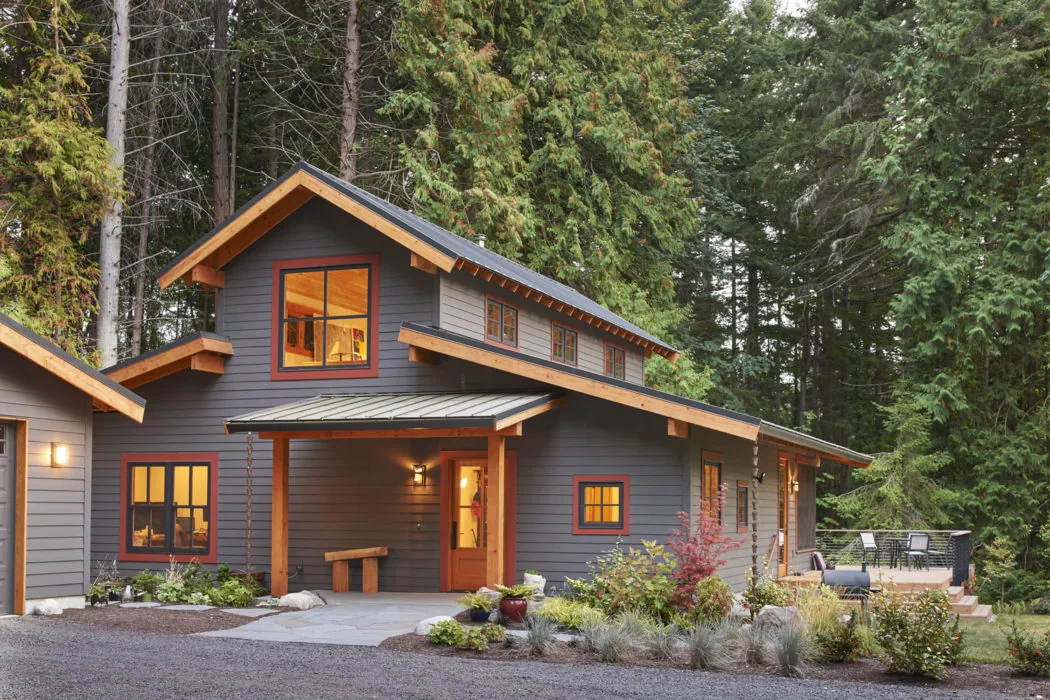 Bainbridge Island Farmhouse features a wraparound porch, wood window trim, and metal railing in a forest setting.