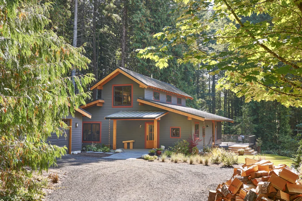 Bainbridge Island Farmhouse by CTA Design Builders features gray wood siding and a metal gable roof framed by tall trees.