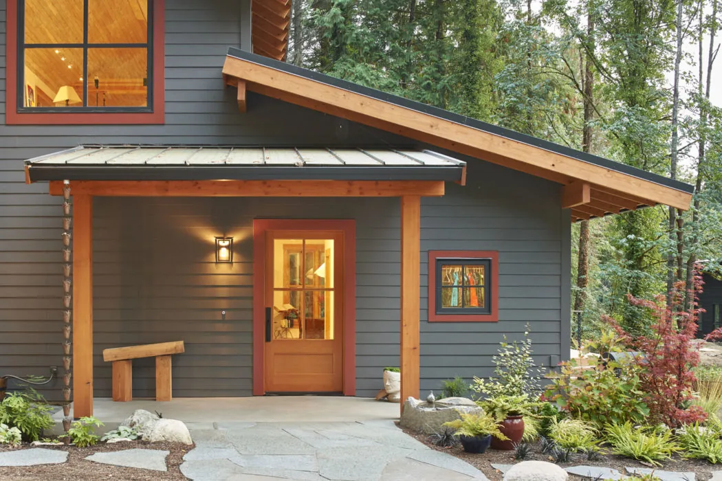 The house entry features wood-trimmed windows and door with a metal roof over the porch, set against forest greenery.