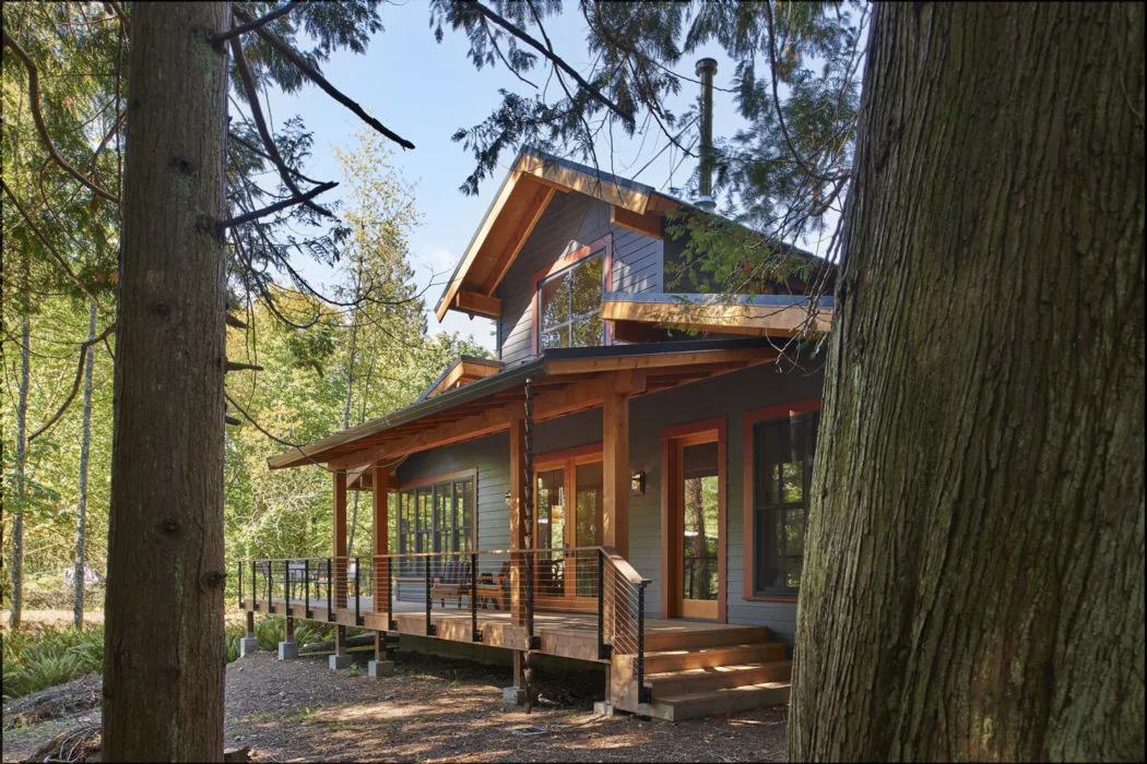Bainbridge Island Farmhouse features wood siding, exposed beams, and a wraparound porch in a wooded setting.