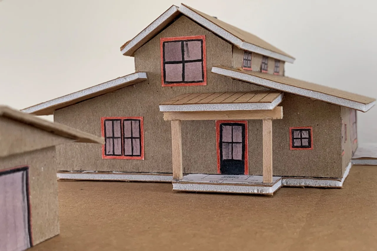 Handmade cardboard model of a farmhouse design with gable roofs and a covered front porch.