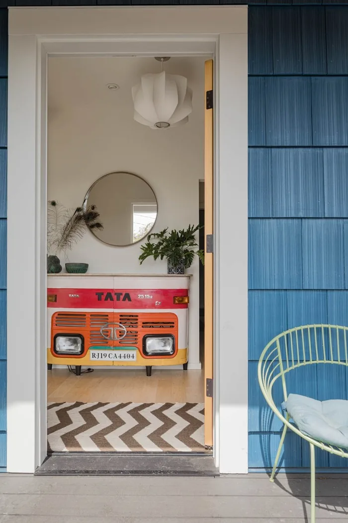View through a doorway into a room with a colorful vintage vehicle front repurposed as a console table beneath a round mirror and modern pet