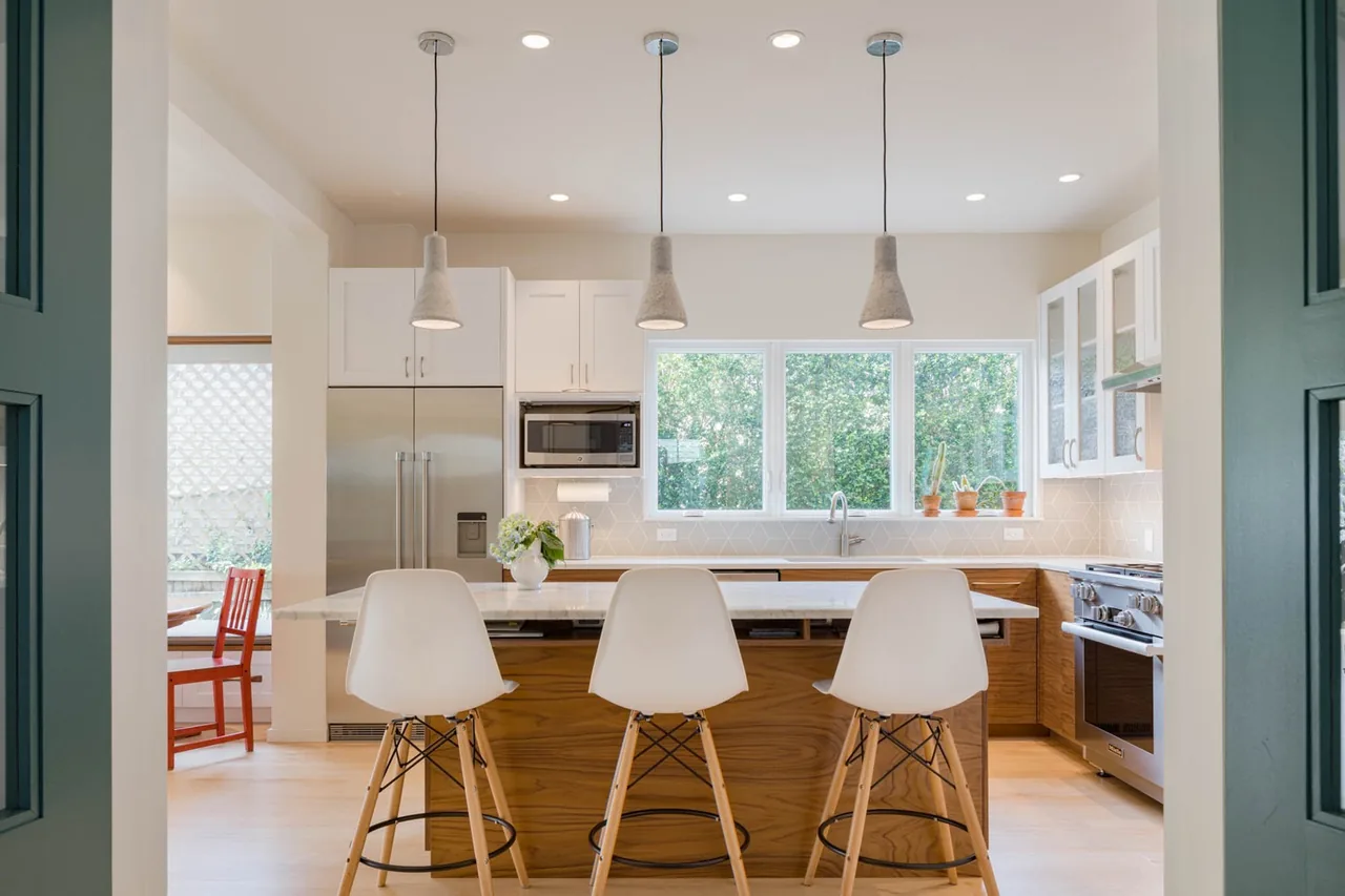 A kitchen featuring wood lower cabinets, white upper cabinets, three pendant lights above a marble island, and large windows overlooking greenery.