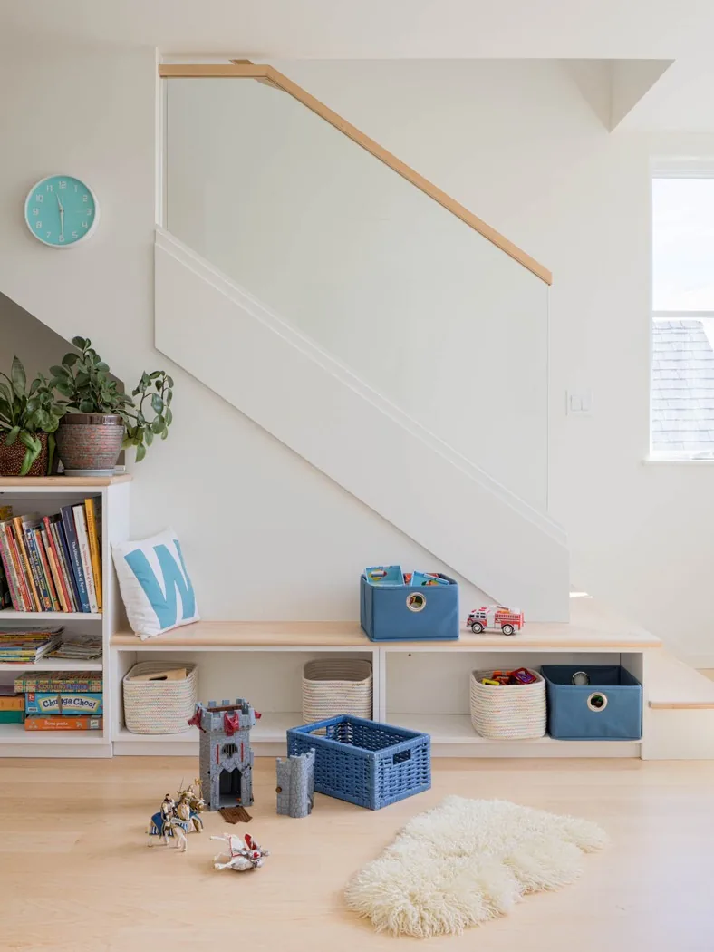 Play area beneath stairs features built-in shelving with baskets and a bench, illuminated by natural light from a nearby window.