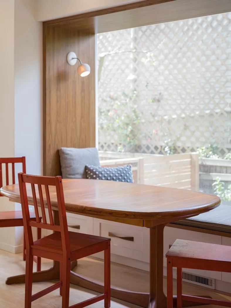 Dining nook features a wooden oval table, red chairs, built-in bench, and a wall-mounted sconce with a window overlooking a privacy lattice.