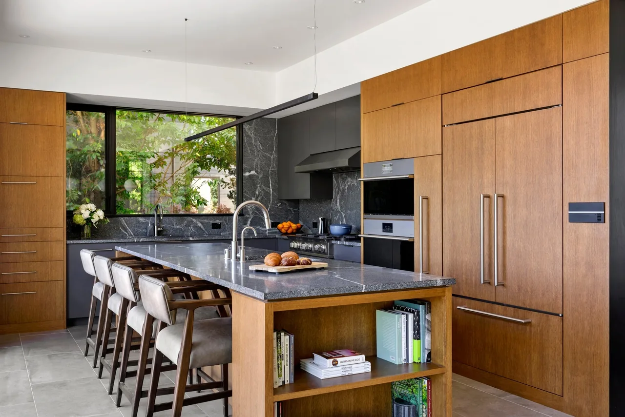 Kitchen features wood cabinets, a gray stone island with built-in shelving, and large black-framed windows.