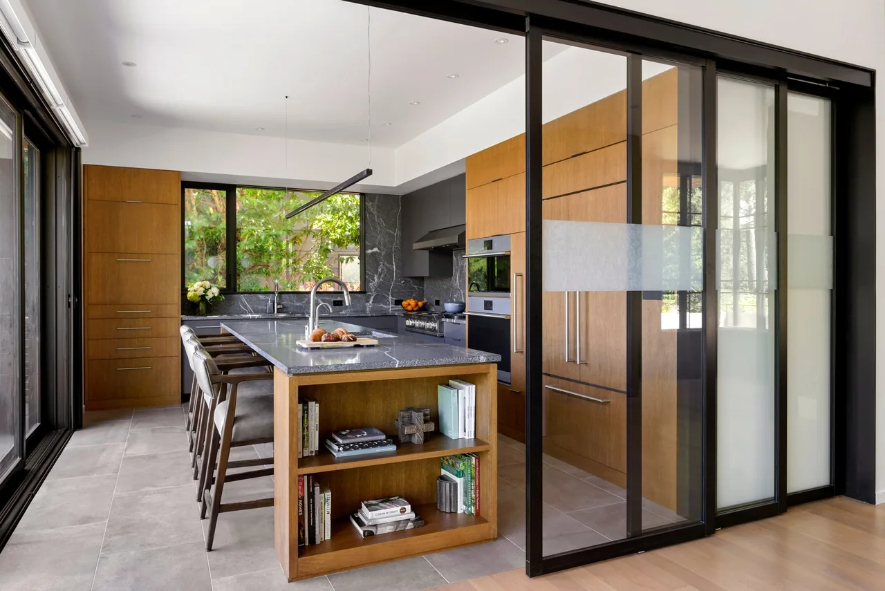 The Sandpoint kitchen by Sticklab Architecture features wood cabinetry with a dark stone island and black window frames.