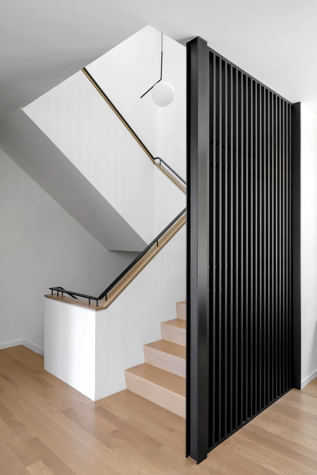 Staircase features light wood treads, black metal handrail, and a black vertical slat divider with a round pendant light above.