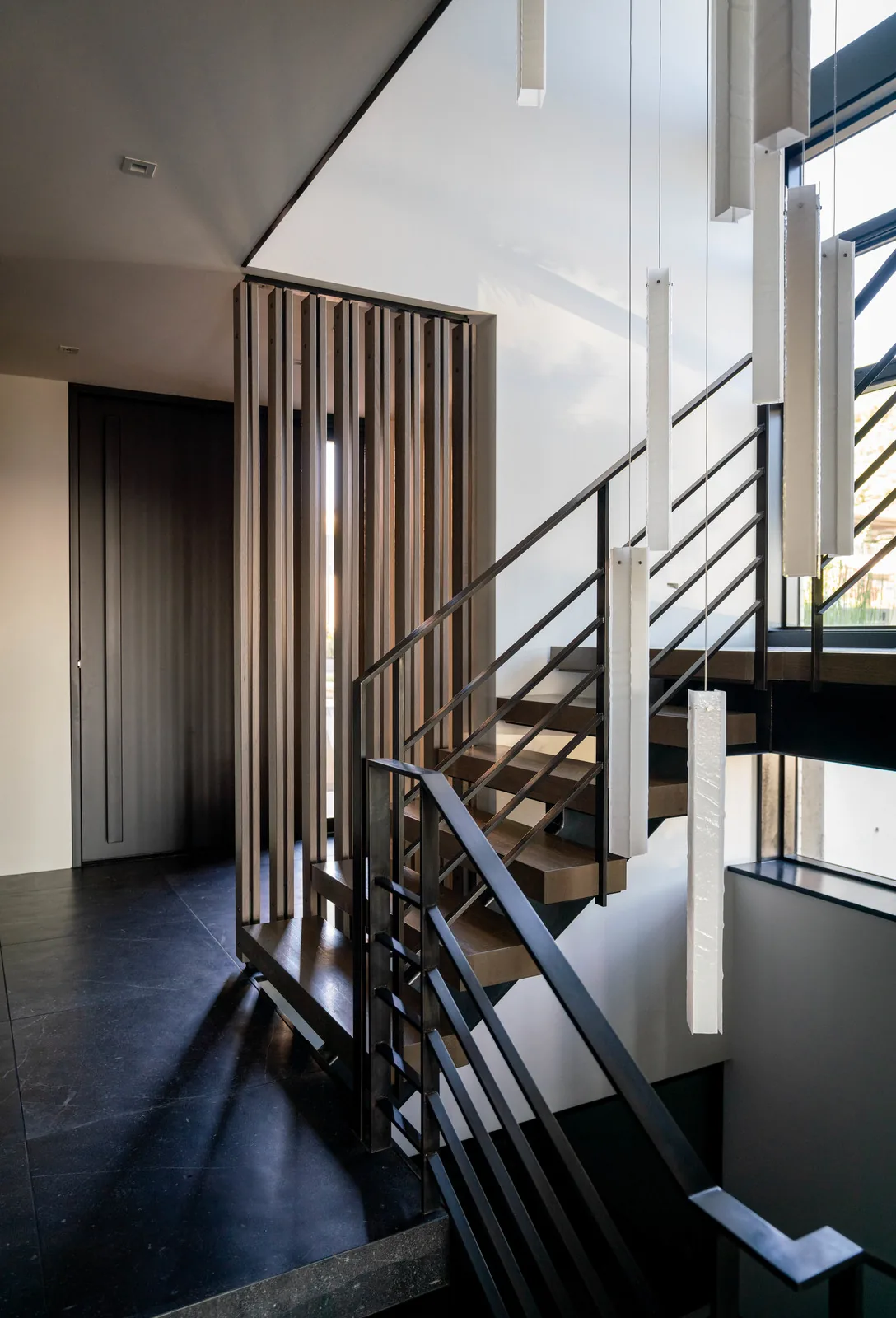 Modern stairwell featuring wooden treads, black metal railing, and vertical wooden slats next to a large window with hanging white pendant lights.