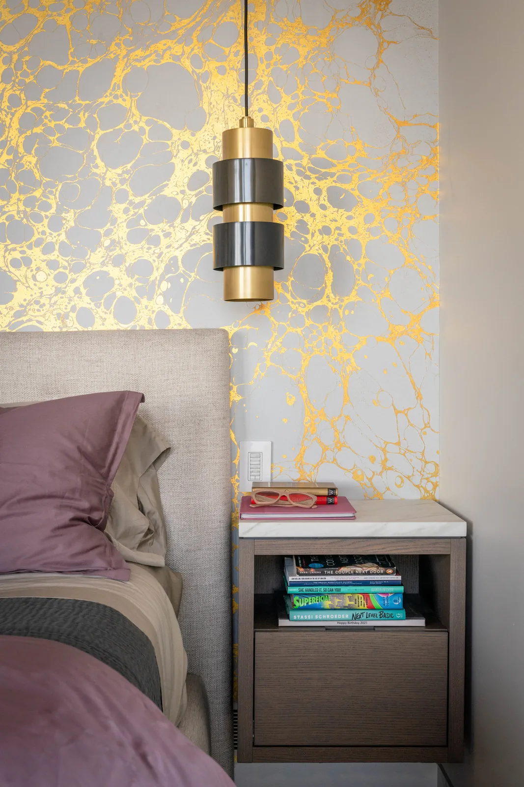 Bedroom corner with beige upholstered headboard, wooden nightstand, and a black and gold pendant light in front of metallic gold patterned wall.