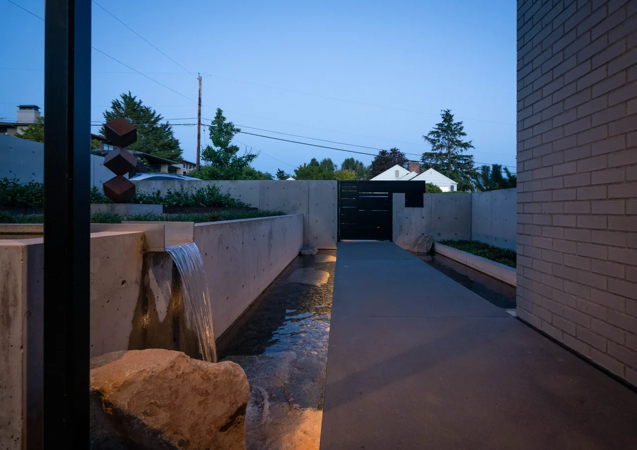 Outdoor pathway over a reflecting pool with water flowing from a concrete wall toward a black metal gate.