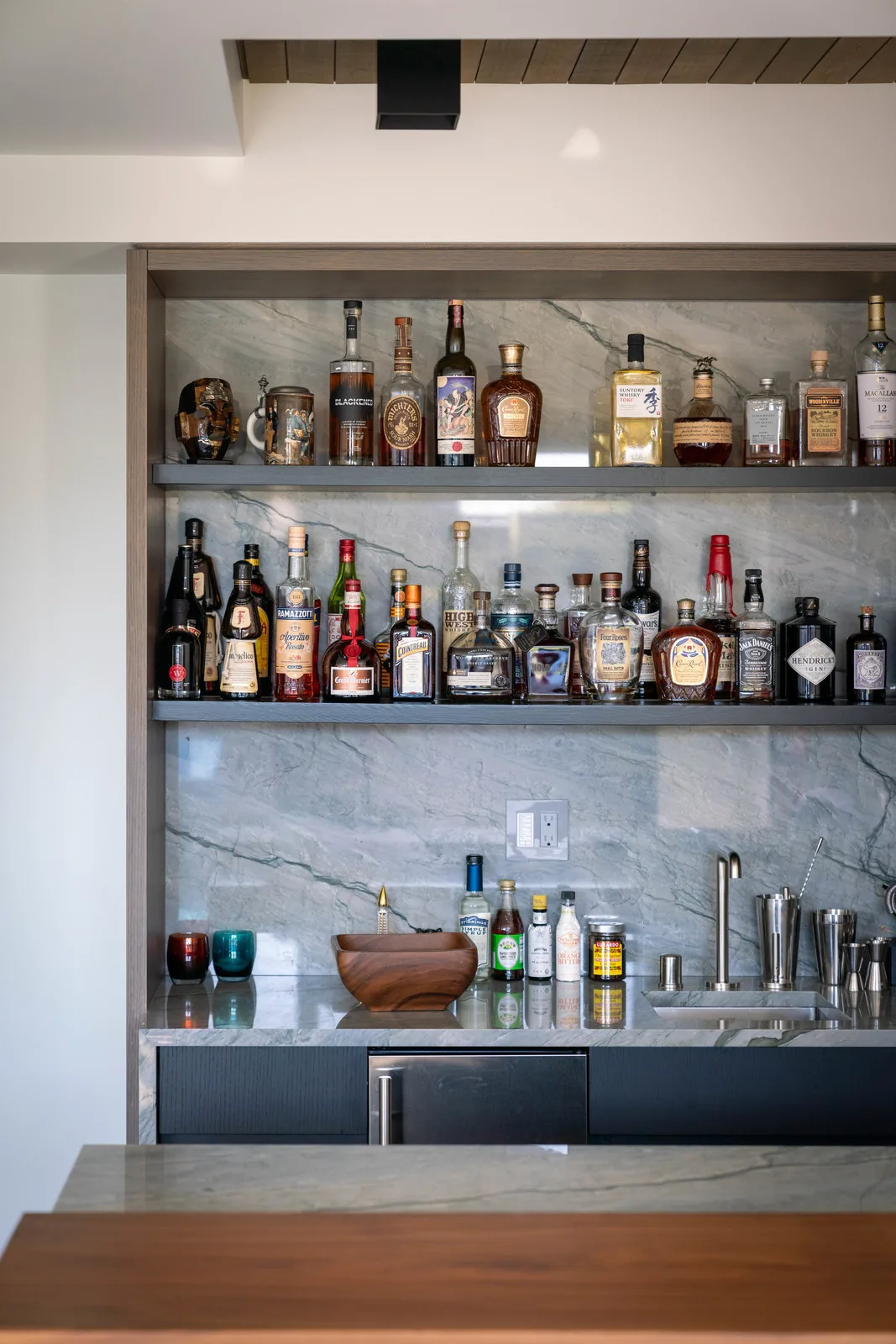 Home bar with marble backsplash, two shelves of liquor bottles, and a stainless steel sink with barware.