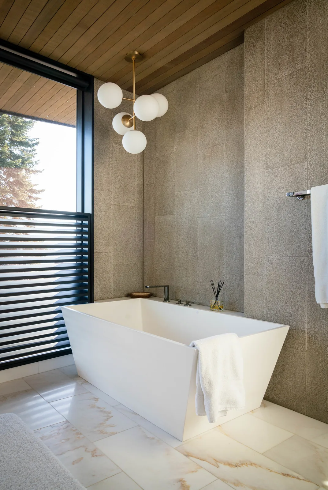A minimalist bathroom features a white freestanding tub beside a large window with black slatted privacy screen.
