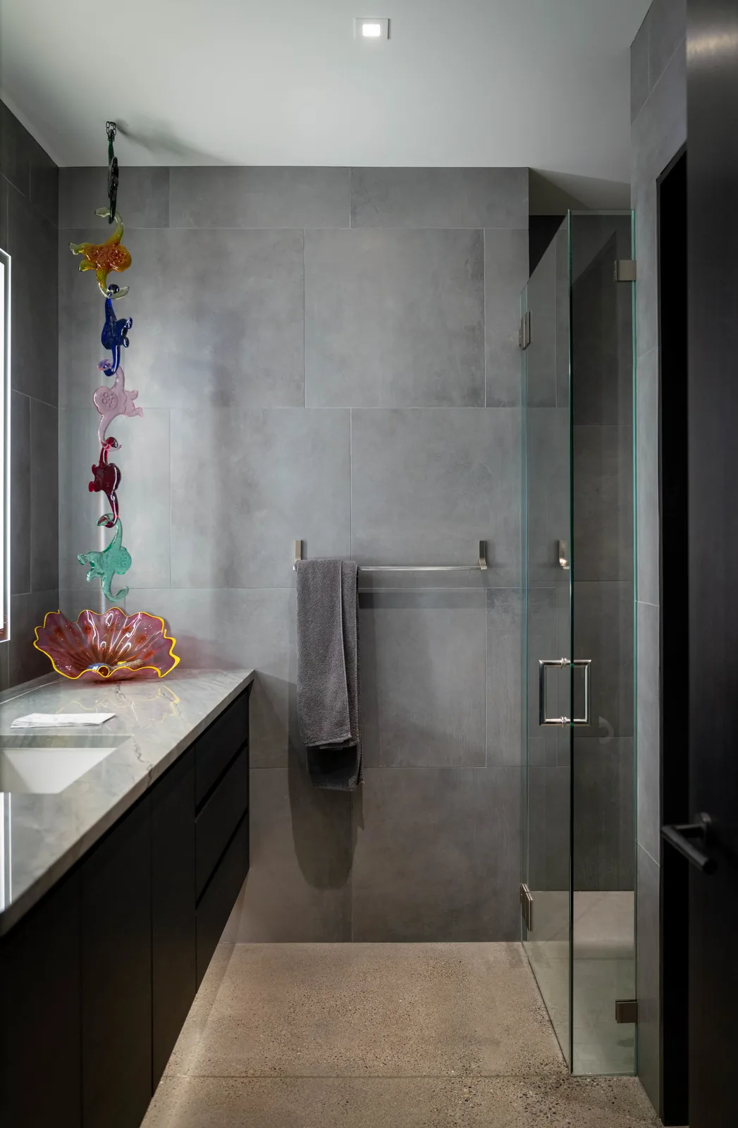 Bathroom featuring textured gray tiles, a glass shower, and a dark vanity with a decorative glass artwork on the countertop.