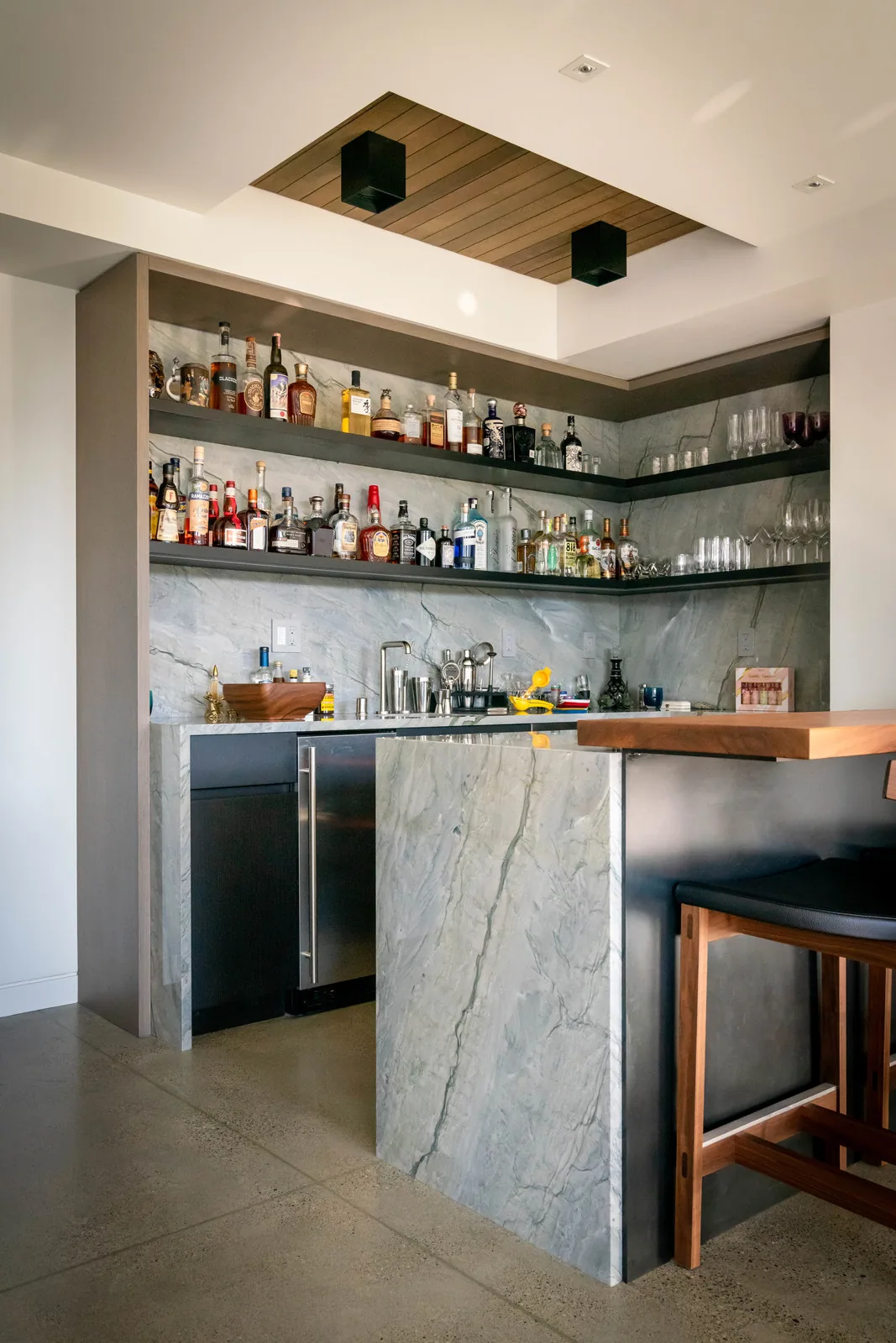 Modern home bar with marble surfaces, open black shelves for bottles, and a small wooden overhead ceiling panel with two black lights.