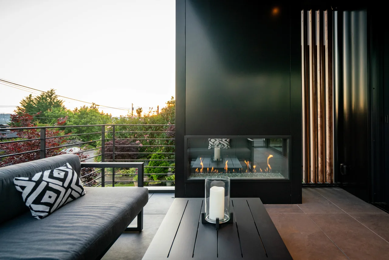 Outdoor patio features a linear fireplace set in a black metal wall and metal railing overlooking greenery at sunset.