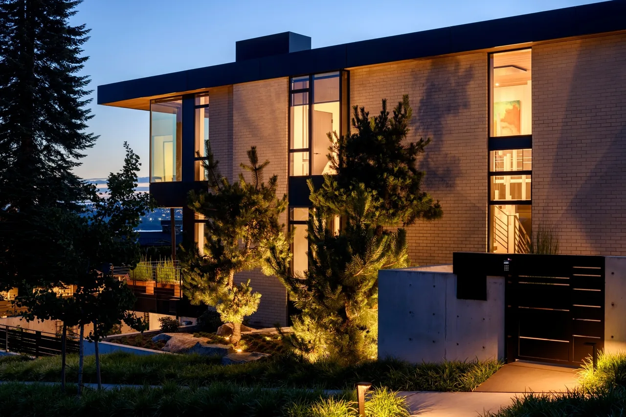 Modern two-story house at dusk with brick facade, large windows, and illuminated landscaping.
