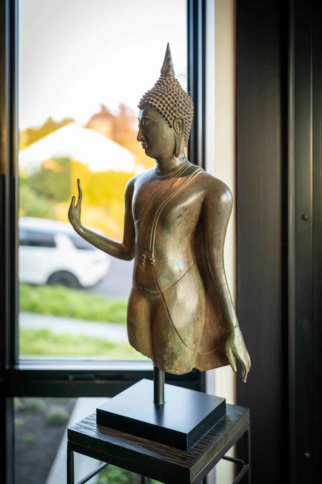 Bronze Buddha statue mounted on a black stand by a large window showing outdoor greenery and a parked white vehicle.