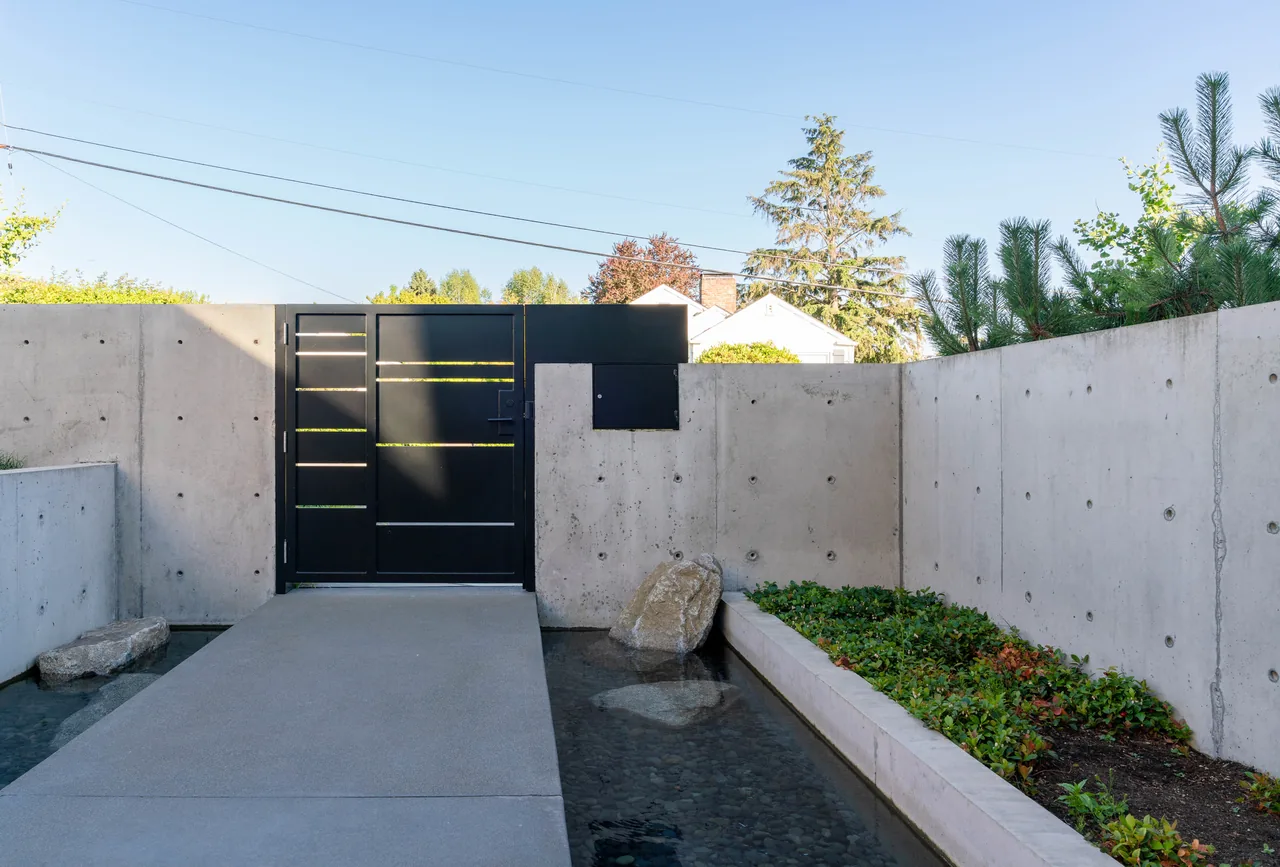 Entry gate with black metal door within a concrete wall, flanked by water features and planter beds at West of Market project.