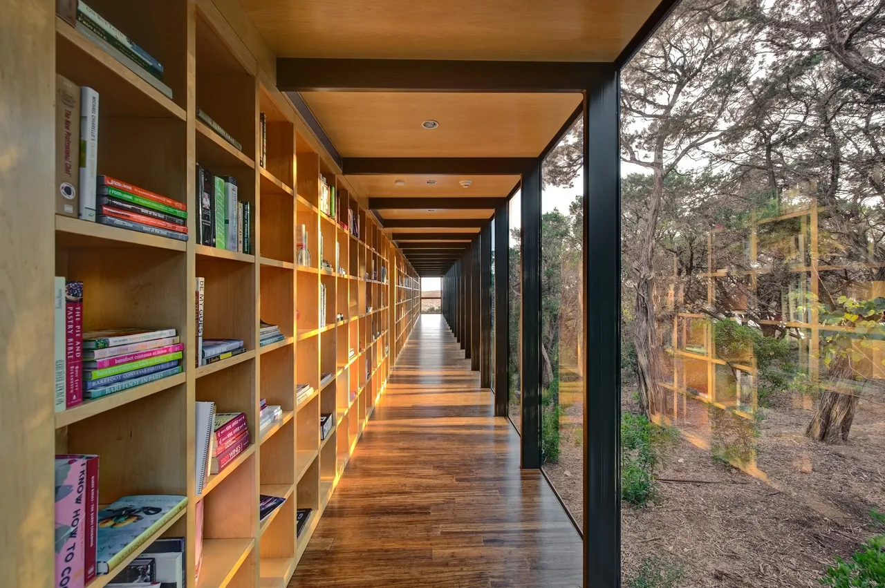 The Wimberley Residence features a hallway with built-in wooden bookshelves and floor-to-ceiling glass windows facing a forest setting.