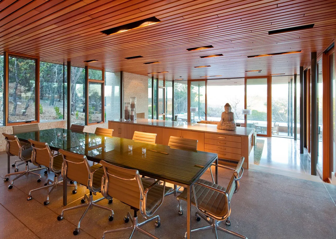 The Wimberley Residence features a spacious glass-walled meeting room with a wood slat ceiling and a long conference table.