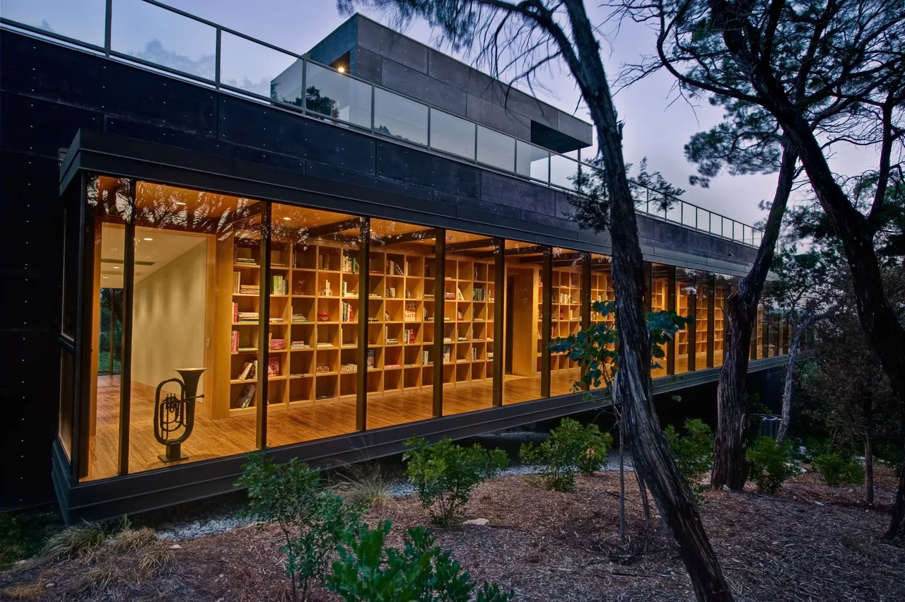 Wimberley Residence shows a library with wood shelving visible through large floor-to-ceiling windows at dusk.