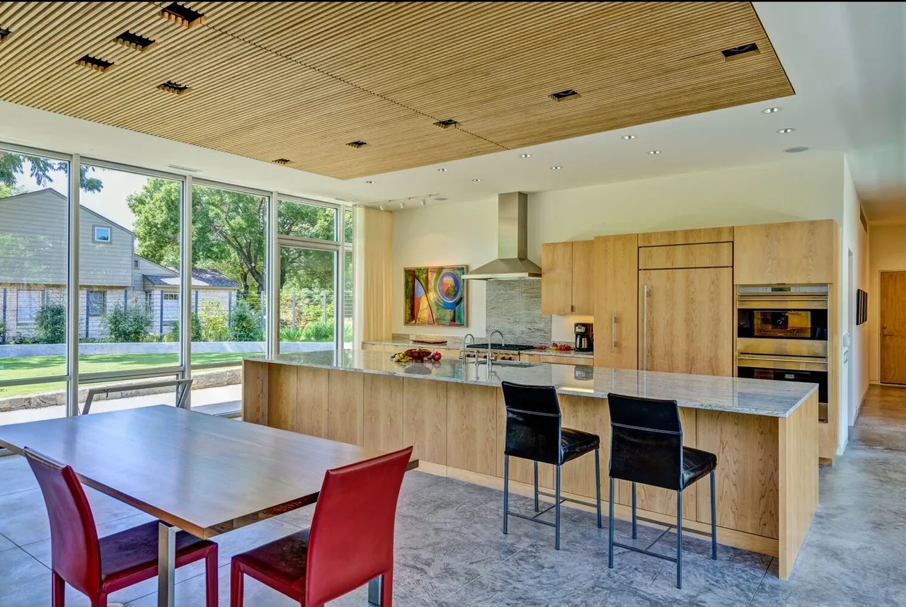 Pomona Residence kitchen features wood cabinetry, stone countertops, and floor-to-ceiling glass windows with a view of the outside lawn.