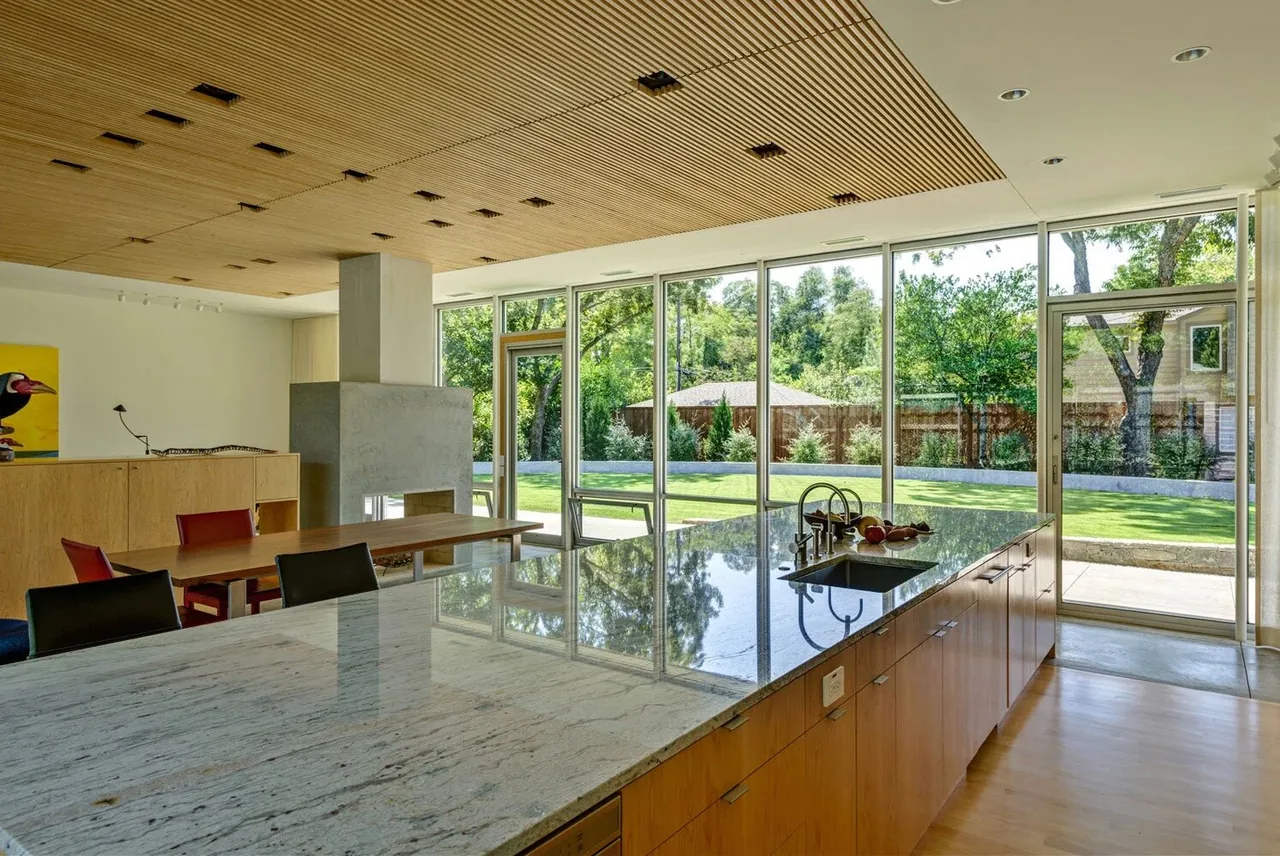 Pomona Residence kitchen features a large marble island and wood slatted ceiling with expansive glass walls to the backyard.