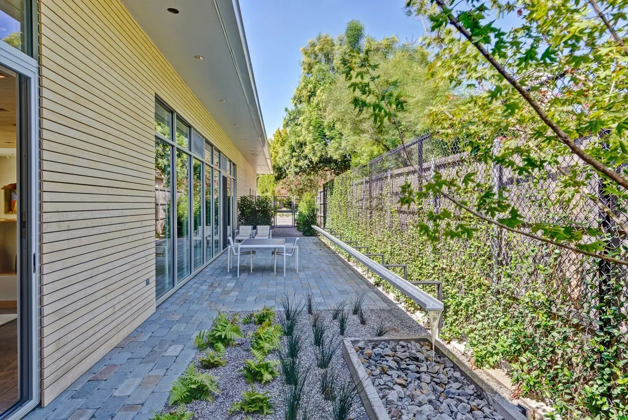 Outdoor side patio with wood siding, floor-to-ceiling windows, paved stone floor, and gravel landscaping with plants and a chain-link fence.