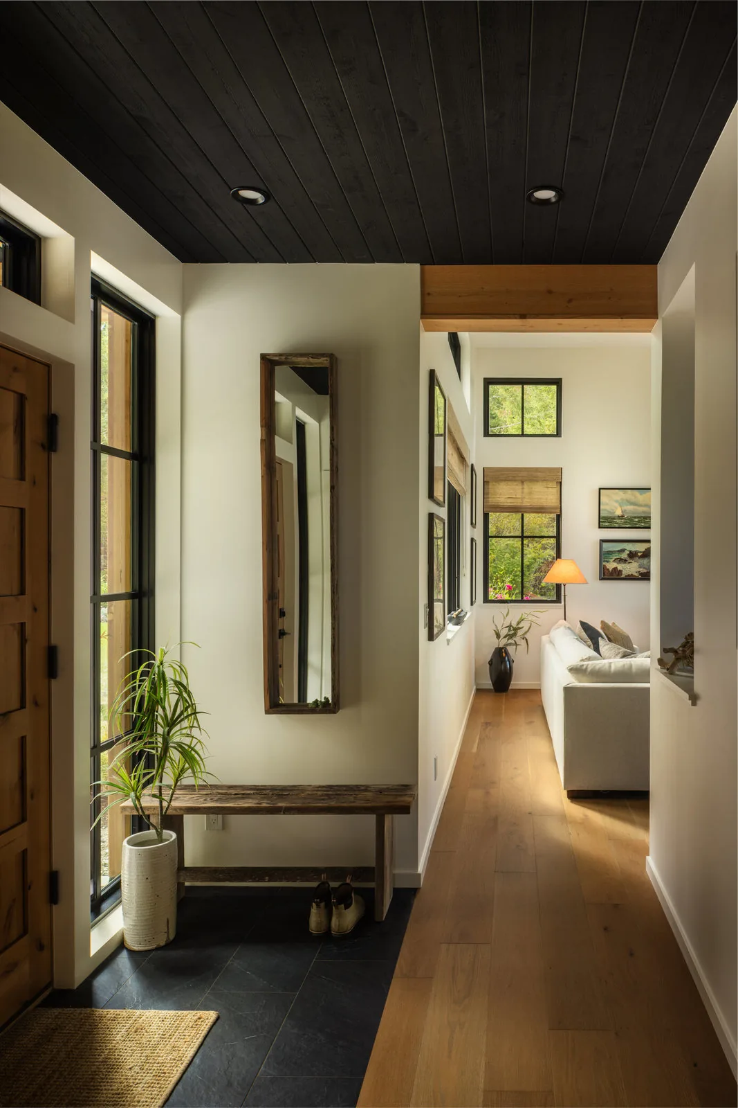 Entry hallway showing a black wood ceiling, wooden bench, mirror, and light-filled living room beyond with framed windows.