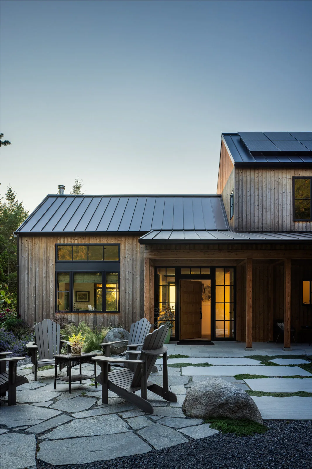 The Fisherman Bay Residence features a stone patio with Adirondack chairs and wood siding under a metal roof with solar panels.