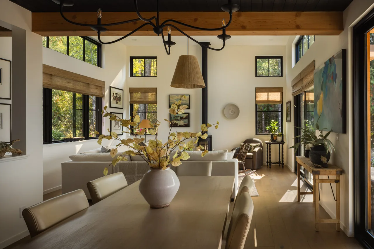Open dining and living room with high ceilings, black-framed windows, and wood flooring, illuminated by natural light.