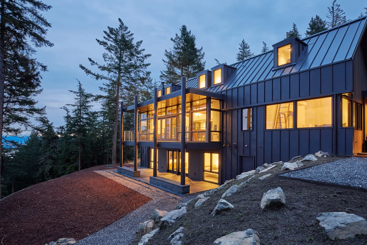 Buck Mountain Residence shows black steel cladding with dormer windows and large floor-to-ceiling glass walls overlooking a forest setting.