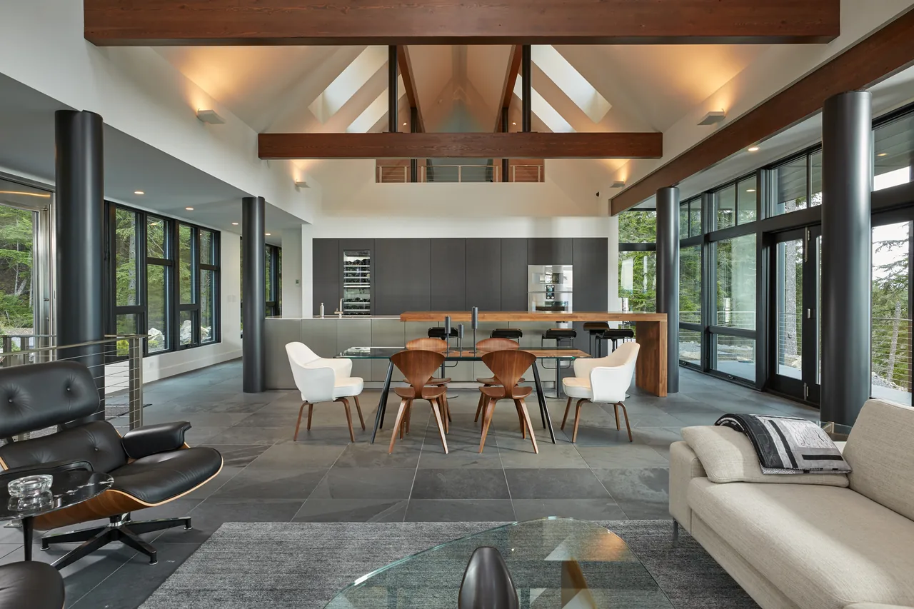 Open living and dining area with vaulted ceiling, wood beams, large black-framed windows, and modern kitchen at Buck Mountain Residence.