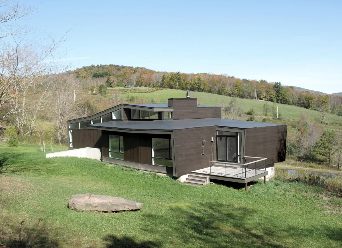 Modern hilltop house with dark wood siding and a large deck surrounded by natural landscape and hills.