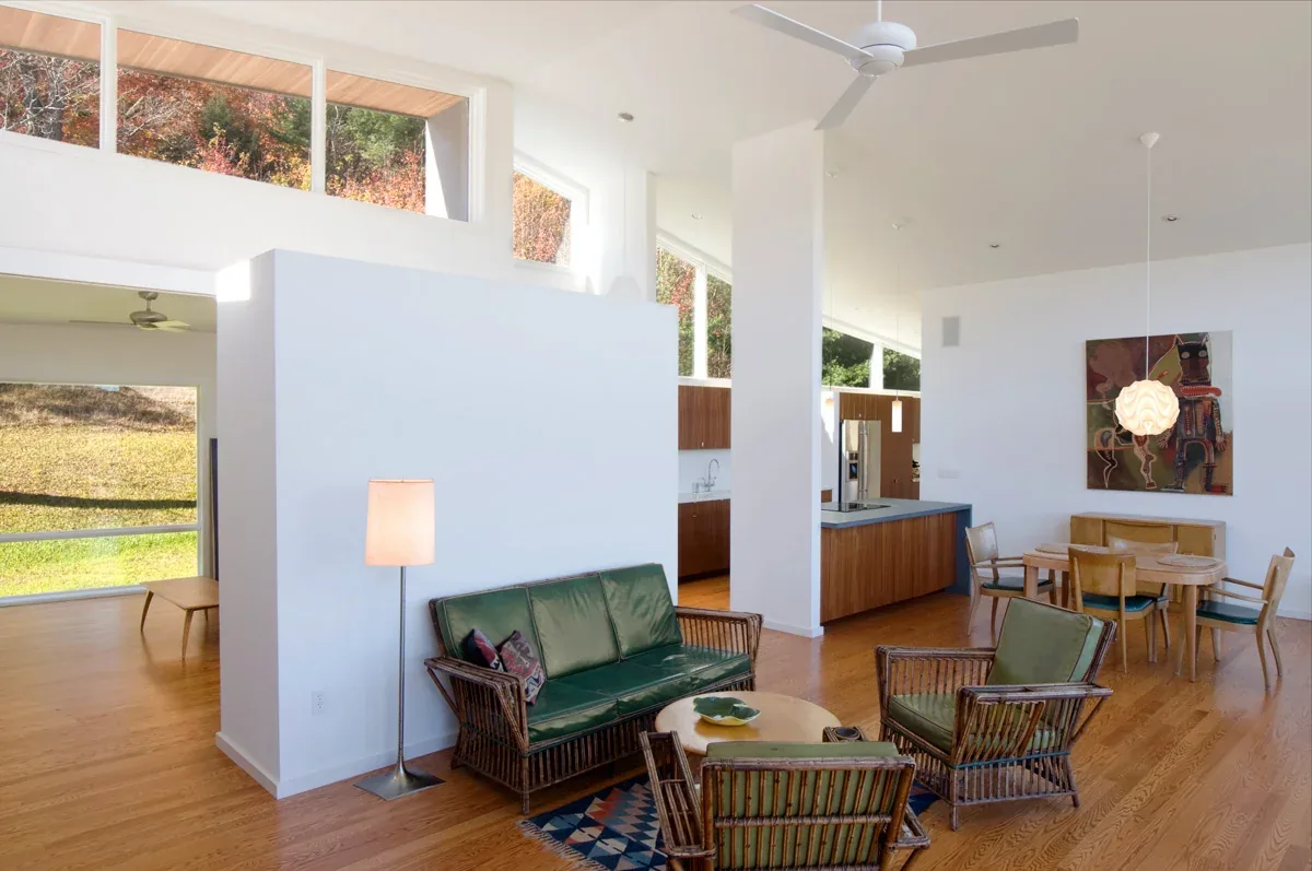 Living and dining area with rattan furniture, wood flooring, and tall clerestory windows overlooking natural landscaping at Quail Ridge House.