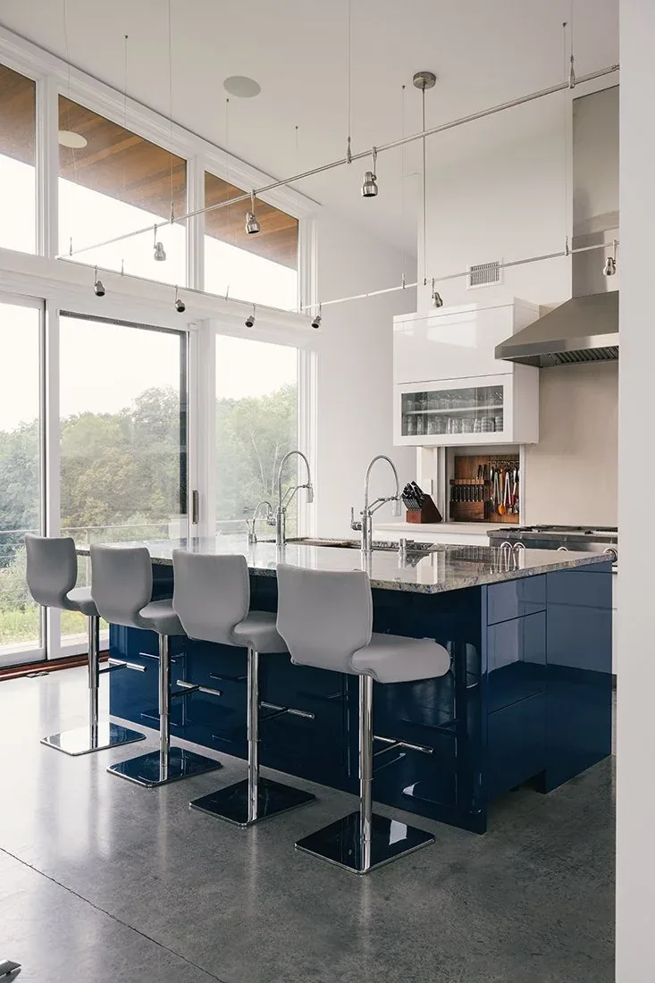 Kitchen with a navy blue island, four gray barstools, and floor-to-ceiling windows overlooking trees.